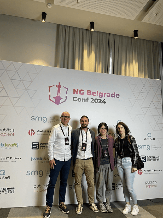 Global IT Factory team stands in front of a white wall with pink geometric patterns and logos at the NG Belgrade Conf 2024, proudly wearing conference badges. The backdrop features the prominent logo of event sponsor Global IT Factory, adding to the vibrant atmosphere.