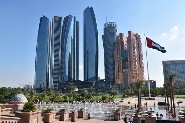 Skyline of Abu Dhabi featuring modern high-rise buildings, a prominent UAE flag, and a foreground with fountains and greenery—exemplifying an environment where advanced infrastructure meets the goals of UAE curriculum management in fostering global standards.